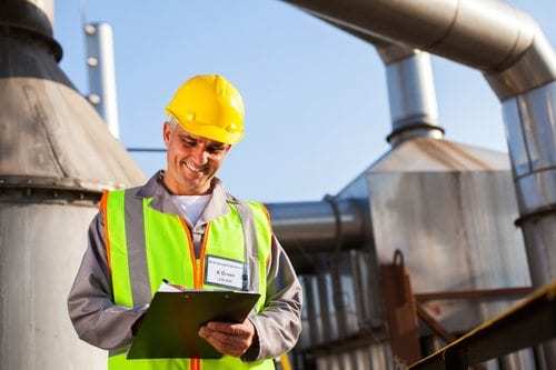 smiling man wearing yellow vest and hard hat writing on clipboard